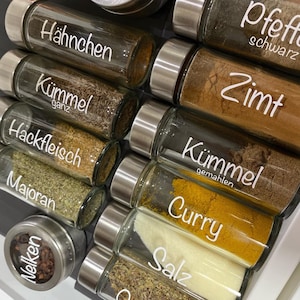 Könnte beinhalten: Eine Sammlung von Gewürzgläsern mit silbernen Deckeln, ordentlich in einer Schublade angeordnet. Jedes Glas ist mit weißem Text beschriftet, darunter "Hähnchen", "Kümmel", "Hackfleisch", "Majoran", "Nelken", "Pfeffer", "Zimt", "Curry" und "Salz".