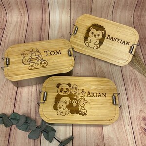 Könnte beinhalten: Drei rechteckige Bambus-Lunchboxen mit eingravierten Tiermotiven und Namen. Eine zeigt ein Eichhörnchen mit einem Fußball und dem Namen "TOM". Eine andere hat einen Igel und den Namen "BASTIAN". Die dritte hat einen Panda und andere Tiere mit dem Namen "ARIAN".