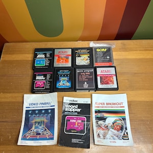 Può includere: Una collezione di cartucce di giochi Atari vintage e manuali di istruzioni. Le cartucce presentano titoli come "Space Jockey", "Word Zapper" e "Indy 500". I manuali includono "Video Pinball" e "Super Breakout", con grafica retrò.