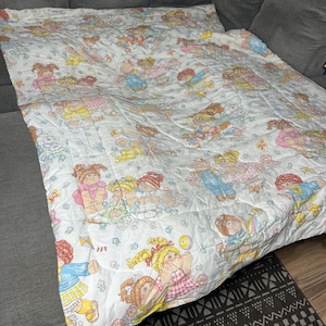 May include: A white quilted blanket featuring a repeating pattern of Cabbage Patch Kids dolls in various outfits, including dresses, overalls, and shirts, accented with small colorful flowers, butterflies, and kites printed across the fabric surface.