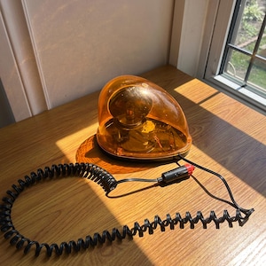 Puede incluir: Una luz de emergencia ámbar con un cable en espiral negro y un enchufe con punta roja. La luz tiene una cubierta redondeada y transparente. El cable en espiral se extiende desde la luz, que está sobre una superficie de madera.