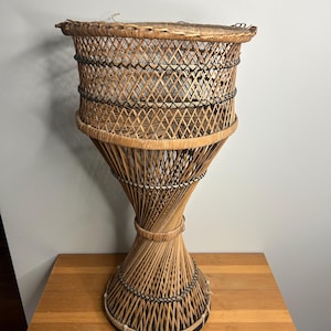 Puede incluir: Un soporte para plantas de mimbre vintage y alto. El soporte presenta una parte superior de cesta tejida, una sección media con diseño de diamantes y una base torcida y acampanada. Los tonos marrones naturales del mimbre se acentúan con detalles negros. Está colocado sobre una superficie de madera.