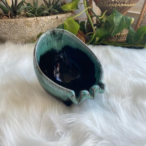 Puede incluir: Un cenicero de cerámica de color verde azulado y negro con un borde ondulado. El cenicero está sobre una superficie blanca y esponjosa.