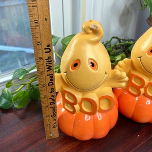 May include: Two vintage Halloween decorations shaped like pumpkins with ghost faces and the word "BOO" in orange letters. The pumpkins are orange and yellow and have a glossy finish.