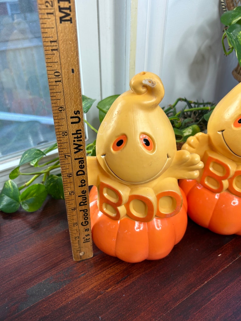 May include: Two vintage Halloween decorations shaped like pumpkins with ghost faces and the word "BOO" in orange letters. The pumpkins are orange and yellow and have a glossy finish.