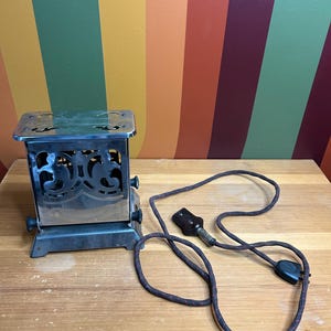 Puede incluir: Una tostadora vintage de plata con un diseño decorativo en la parte delantera. La tostadora tiene un cable negro con un enchufe en cada extremo.