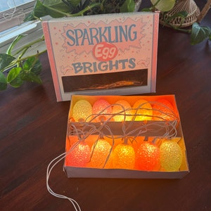 Puede incluir: Una caja de 10 luces de huevo pastel con un cable blanco. La caja es rosa y blanca y dice "Sparkling Egg Brights".