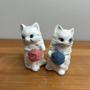 May include: Two white ceramic cat figurines, each holding a ball of yarn. One cat holds a pink yarn ball, the other a blue one. The cats have blue eyes and black whiskers. The figurines are on a wooden surface.