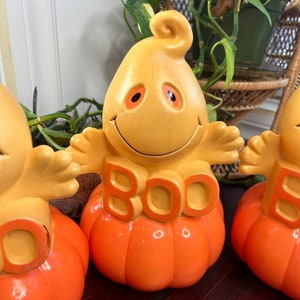 May include: Three orange and yellow ceramic Halloween decorations shaped like pumpkins with ghosts sitting on top. The ghosts have orange eyes and say "BOO" on the front.