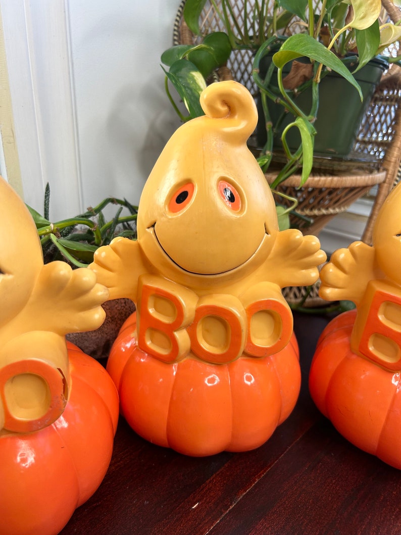 May include: Three orange and yellow ceramic Halloween decorations shaped like pumpkins with ghosts sitting on top. The ghosts have orange eyes and say "BOO" on the front.