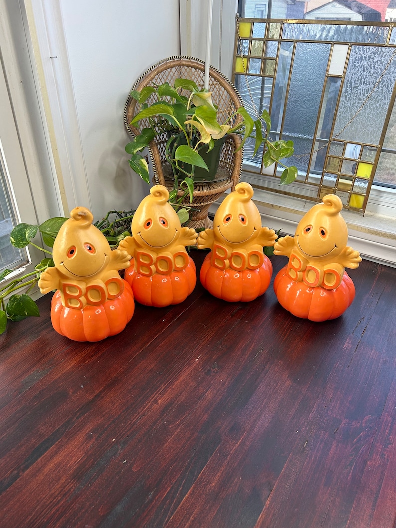 May include: Four ceramic pumpkin figurines with ghost faces and the word "BOO" on the front. The pumpkins are orange and yellow and are sitting on a wooden surface.