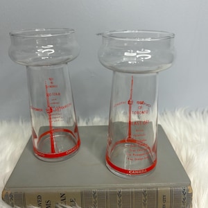 May include: Two clear glass tumblers with a red rim and red text. The text includes "TORONTO" and "CANADA". The tumblers have a unique shape, with a wide top and a narrower base. The glasses are on a gray book.