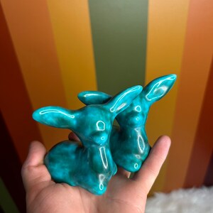 May include: Two turquoise ceramic bunny figurines. The bunnies are in a sitting position with their ears up. The ceramic has a glossy finish. The background is a colourful striped pattern of orange, yellow, and green.