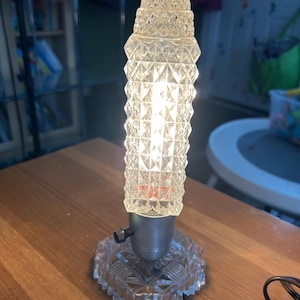 Könnte beinhalten: Eine Tischlampe aus klarem Glas mit einem rautenförmigen Muster. Die Lampe hat einen silbernen Sockel und ein klares Kabel.