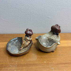 Peut inclure: Deux figurines de grenouilles en céramique. L'une est assise sur un plat en forme de feuille, l'autre dans un petit bol. Les deux grenouilles ont des têtes marron et portent des nœuds papillon. Le plat et le bol sont de couleurs neutres.