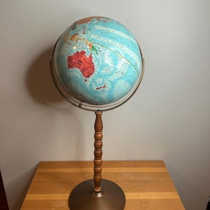 Może przedstawiać: Zabytkowy globus z jasnoniebieskim oceanem i kolorowymi kontynentami, zamontowany na ciemnobrązowym metalowym stojaku z drewnianą kolumną. Globus jest podtrzymywany przez metalowy południk i stoi na okrągłej podstawie.