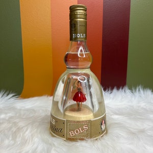 Puede incluir: Una botella de licor de oro Bols bailarina vintage. La botella de vidrio transparente tiene una tapa dorada y una figura de bailarina en el interior. La etiqueta dice "Bols" y "Gold Liqueur". La botella está sobre una superficie blanca y esponjosa.