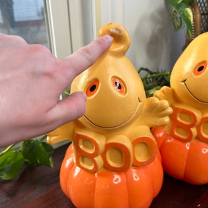 May include: Two orange and yellow ceramic pumpkin figurines with ghost faces and the word "BOO" on the front.