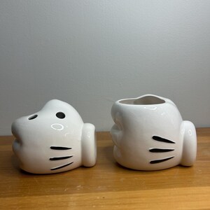 May include: Two white ceramic Mickey Mouse glove-shaped containers. One has a hole on top and the other has two holes. Both have black details, including three lines on the palm and two black dots. They are on a wooden surface.