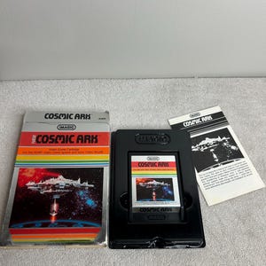Puede incluir: Un cartucho de videojuego "Cosmic Ark" vintage para el sistema Atari. La caja presenta un gráfico de nave espacial con el título del juego en rojo y blanco. El cartucho está en una caja negra con un manual de instrucciones a juego.