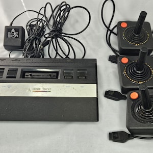 Puede incluir: Una consola de videojuegos Atari 2600 vintage con tres joysticks y un adaptador de corriente. La consola es negra con una franja plateada y el logotipo de Atari. Los joysticks son negros con botones naranjas.