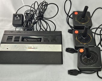 Puede incluir: Una consola de videojuegos Atari 2600 vintage con tres joysticks y un adaptador de corriente. La consola es negra con una franja plateada y el logotipo de Atari. Los joysticks son negros con botones naranjas.