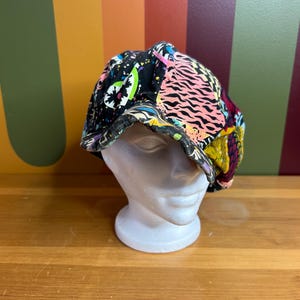 Puede incluir: Una gorra de estilo newsboy con estampado negro, verde, rosa y amarillo. La gorra tiene una variedad de diseños abstractos y está hecha de un tejido de algodón suave.