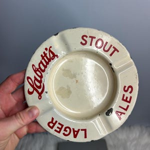 May include: A vintage off-white ashtray with red lettering. The ashtray features the Labatt's logo and the words STOUT, ALES, and LAGER. The ashtray has a circular design with a small indentation for cigarettes.