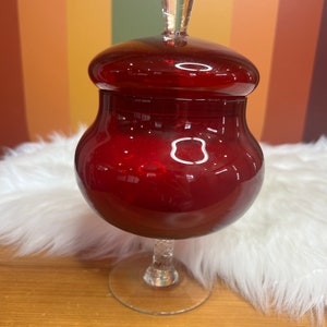 Puede incluir: Un tarro de dulces de vidrio rojo con una tapa de vidrio transparente y una base de pedestal de vidrio transparente.