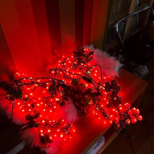 May include: Red Christmas lights shaped like grapes with green leaves, draped over a white fluffy surface and a wooden table.