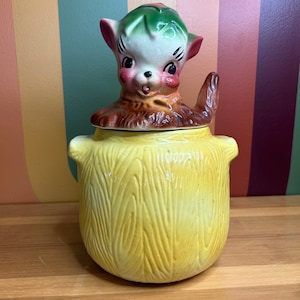 May include: A yellow ceramic cookie jar with a squirrel lid. The squirrel is brown and green with a white face and black eyes. The jar has a wood grain pattern.