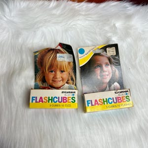 May include: Two boxes of Sylvania Blue Dot Flashcubes. Each box contains 3 cubes, which provide 12 flashes. The boxes feature a blue dot logo and a photo of a young girl.