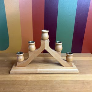 May include: A wooden candle holder with five candle cups. The holder is shaped like a triangle with a base and two sides that slope upwards. The candle cups are all the same size and are evenly spaced along the top of the holder.