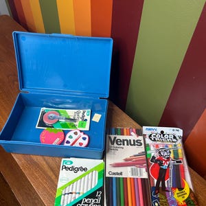 May include: A blue art supply box containing art materials. The box holds a pack of Venus 12 Colouring Pencils, a pack of Pedigree 12 pencil crayons, and a pack of Marvy Color Tricks markers. Also included are erasers and scissors.