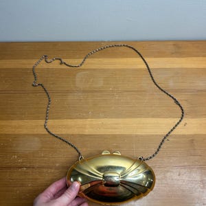 Könnte beinhalten: Eine goldfarbene, ovale Clutch mit einem schleifenförmigen Verschluss und einem silbernen Kettenriemen. Die Clutch wird von einer Hand gehalten, der Hintergrund ist eine Holzoberfläche.