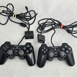 May include: Two black Sony PlayStation controllers with attached cords. Each controller has a directional pad, action buttons, and analog sticks. The cords are bundled with a yellow rubber band and an orange rubber band.