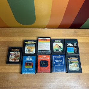 Peut inclure: Une collection de cartouches de jeux Atari vintage, dont "Home Run", "Haunted House", "Atlantis", "Othello", "Oink!", "Spider Fighter", "Keystone Kapers" et "Super Breakout". Les cartouches sont de couleurs variées et présentent des titres et des graphismes de jeux.