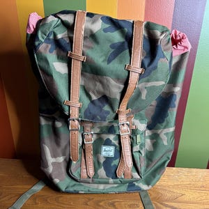 Op de afbeelding: Camouflage rugzak met bruine leren riemen en zilveren gespen. De rugzak heeft een voorvak en verstelbare schouderbanden. De binnenvoering is rood en wit gestreept. De merknaam is zichtbaar.