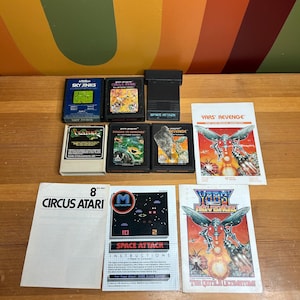 Puede incluir: Una colección de cartuchos y manuales de videojuegos Atari vintage. Los títulos incluyen "Sky Jinks", "Circus Atari", "Space Attack" y "Yars' Revenge". Los cartuchos son de varios colores y los manuales presentan gráficos coloridos.