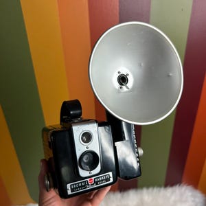 Puede incluir: Una cámara Brownie Hawkeye Flash Model negra con un accesorio de flash plateado. La cámara tiene un círculo rojo con la palabra "Brownie" y las palabras "Hawkeye Flash Model" impresas en ella.