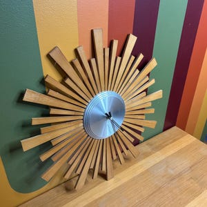 May include: A mid-century modern wall clock with a sunburst design. The clock has a silver-coloured face and wooden rays radiating outwards. The clock is displayed on a wooden surface against a colourful striped background.