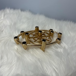 May include: A gold-coloured metal candle holder with a circular design. The holder has multiple cylindrical openings for candles, with black interiors. The design includes a central wheel-like structure and an outer ring.