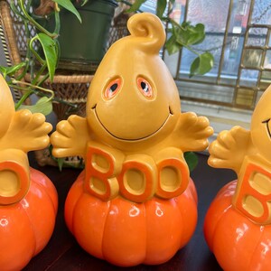 May include: Three ceramic Halloween decorations featuring a ghost with a smiling face and the word "BOO" on a pumpkin. The ghosts are yellow with orange eyes and the pumpkins are orange.