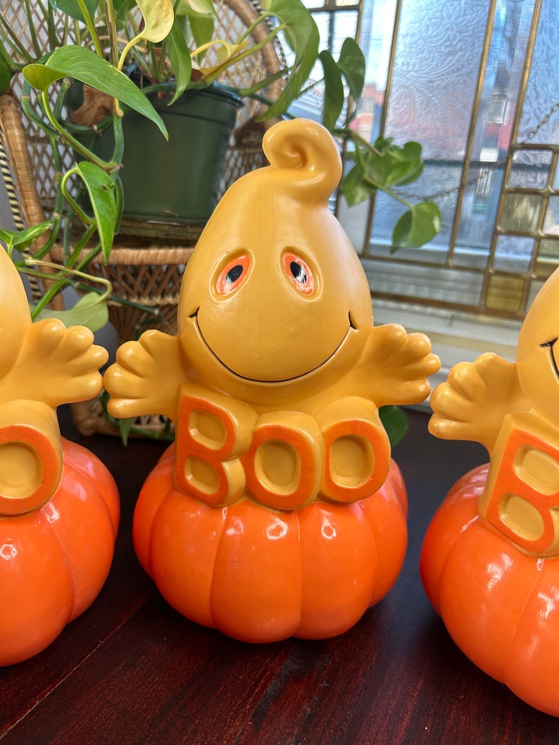 May include: Three ceramic Halloween decorations featuring a ghost with a smiling face and the word "BOO" on a pumpkin. The ghosts are yellow with orange eyes and the pumpkins are orange.