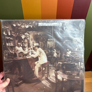 May include: A vintage vinyl record album cover featuring a black and white photo of a man in a white suit sitting at a bar with other people in the background. The album cover is titled "Led Zeppelin - "In Through The Out Door"/Cover"
