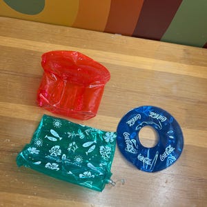 May include: Three inflatable pool toys: a red rectangular shape, a green rectangular shape with white designs, and a blue ring with "Coca-Cola" branding. The toys are on a wooden surface.