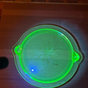 Può includere: Un vassoio rotondo in vetro verde neon con due manici. Il vassoio presenta un motivo floreale inciso al centro. Le parole "MILLER'S GENERAL STORE" sono visibili sullo sfondo.