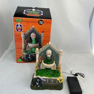 Puede incluir: Una fuente de pueblo espeluznante con un cráneo y slime verde que sale de su boca. La fuente está hecha de resina y tiene una función de iluminación. La fuente forma parte de la colección Spooky Town de Lemax.