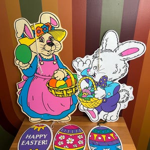 Peut inclure: Décorations sur le thème de Pâques avec deux découpes de lapins et trois ornements en forme d'œufs. Les lapins sont vêtus de tenues colorées et tiennent des paniers et des œufs. Les ornements en forme d'œufs ont des motifs floraux et géométriques.