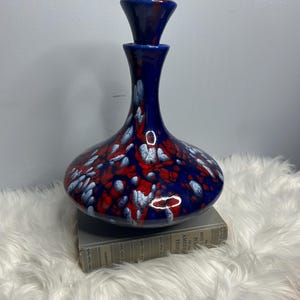 May include: A navy blue ceramic decanter with a matching stopper. The decanter has a bulbous base and a slender neck. It is decorated with red and white speckled accents. The decanter is sitting on a book.